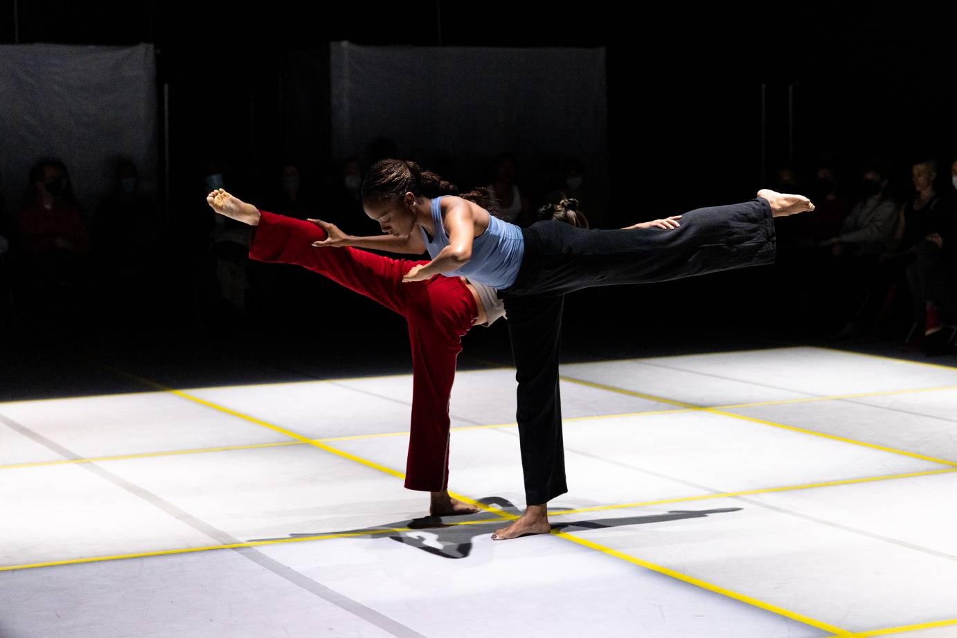 two dancers on a gray stage designed with yellow tape in rectangle shapes extend their legs in arabesque one facing left the other facing right. The hold on to one anothers out streched legs